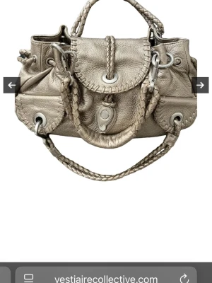 Silver handväska från Ted Lapidus i skinn - Säljer en snygg handväska i metallic silverfärgat skinn. Väskan har flätade handtag och detaljer, stora metalldelar och en unik lockstängning. Insidan har dragkedja och flera fack. Perfekt för dig som gillar statement-accessoarer.köpt från vestariecollective