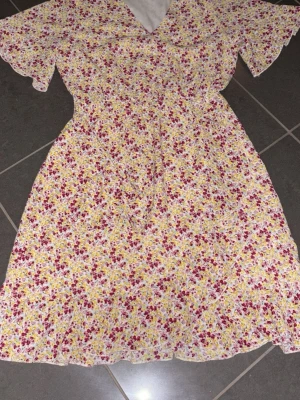 Blommig omlottklänning med volang - Säljer en söt omlottklänning med blommigt mönster i rosa, gul och vit. Klänningen har korta volangärmar, v-ringad hals och volangkant nedtill. Perfekt för sommarens alla tillfällen och har en luftig passform.