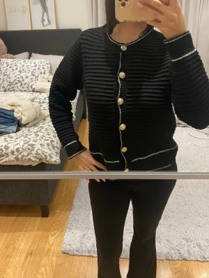 Svart stickad kofta med guldfärgade knappar - Säljer en svart stickad kofta med snygga guldfärgade knappar framtill. Koftan har vita detaljer längs kanterna och muddar, samt ett ribbat mönster som ger en cool struktur. Perfekt för dig som gillar klassisk stil med en twist.