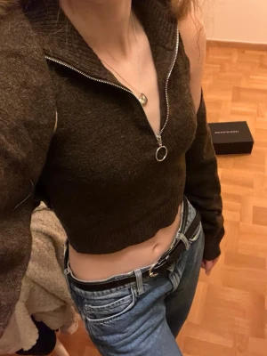 Brun croppad stickad polotopp med zip - Säljer en brun croppad stickad polotopp med lång ärm och stor krage. Toppen har en snygg silverfärgad dragkedja med ringdetalj framtill. Materialet är mjukt och ribbat, perfekt för dig som gillar trendiga och bekväma plagg.