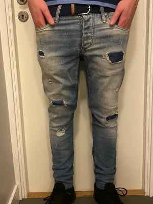 Blå slitna jeans - Säljer ett par blå jeans med slitningar och hål på flera ställen, vilket ger en cool och avslappnad vibe. Modellen är skinny med smal passform och klassisk femficksdesign. Jeansen har ljus tvätt och är gjorda i jeansmaterial med stretch. Orten grish?