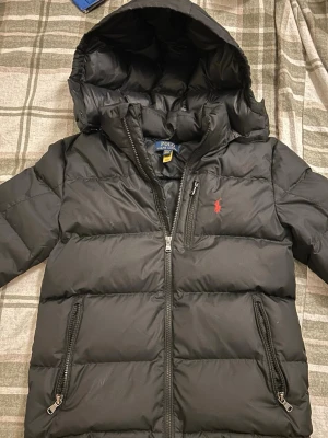 Svart pufferjacka från Polo Ralph Lauren - Svart pufferjacka från Polo Ralph Lauren med huva och två sidofickor med dragkedja. Jackan har ett rött Polo-logo broderat på bröstet och är tillverkad i syntetmaterial. Perfekt för kalla dagar och har en schysst, sportig vibe. Nypris 3300