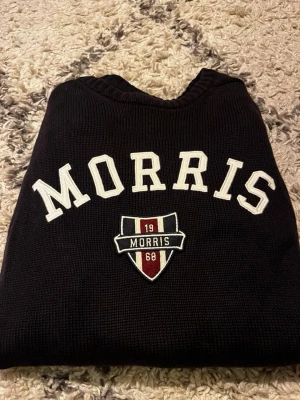 Svart stickad tröja Morris XS Passar som S/M - Svart stickad tröja från Morris i storlek XS. Tröjan har vit broderad text 'MORRIS' och ett snyggt emblem på bröstet med röd, vit och blå detalj. Rund hals och långa ärmar med ribbade muddar. Perfekt för dig som gillar klassisk stil. Tröjan sitter mer som S/M och inte som XS.