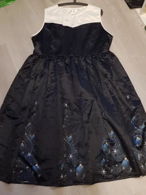 Ny/ Oanvänd Marinblå och vit långklänning Nextmia - Långklänning från Nextmia med vit ärmlös överdel i prickigt tyg och svart glansig kjol. Kjolen har tryck med blå julkulor och stjärnor nertill. Klänningen har dragkedja i sidan och är tillverkad i ett satinliknande material.Kanske något för Nyår!! 😊