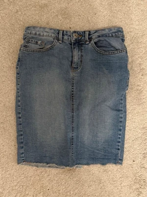 Blå jeanskjol från ONLY, storlek 36 - Säljer en klassisk blå jeanskjol från ONLY i storlek 36. Kjolen har fem fickor, bälteshällor och rå kant nedtill för en cool vibe. Baksidan har två fickor med snygga sömmar och en liten slits. Perfekt att matcha med sneakers eller boots.