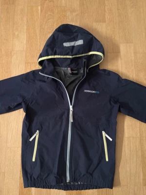Mörkblå vindjacka från Didriksons - Snygg mörkblå vindjacka från Didriksons med huva och ljusgula detaljer. Jackan har två fickor med dragkedja, reflexdetaljer och logga på bröstet och ryggen. Perfekt för blåsiga dagar och har en sportig vibe. Storlek: 10år 140cm