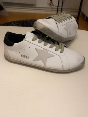 Golden Goose vita sneakers med stjärna - Säljer ett par vita Golden Goose sneakers med grå mockadetaljer och en stor stjärna på sidan. Skorna har grå snörning, svart häl och GGDB-logga i metall. Själv köpta på plick så ingen aning om de är äkta eller inte därför säljer jag för ett schysst pris. Ny skick