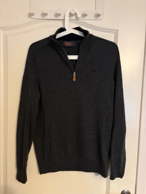 Mörkgrå half zip tröja Morris - Snygg mörkgrå långärmad tröja från Morris med half zip och liten broderad logga på bröstet. Tröjan har en klassisk passform och är tillverkad i mjukt material som passar perfekt för kyliga dagar. Perfekt att bära över en t-shirt eller skjorta.