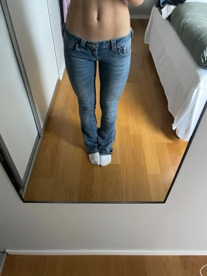 True Religion lågmidjade blå jeans - Säljer ett par blå jeans från True Religion med låg midja och klassisk femficksdesign. Jeansen har en lätt tvättad look, raka ben och synliga sömmar. Märkespatch bak i linningen och snygga detaljer på bakfickorna. Perfekt för dig som gillar en avslappnad stil.