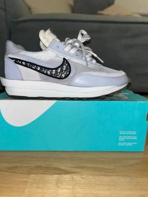 Nike x Sacai sneakers med Dior-mönster - Nike x Sacai sneakers i ljusgrått och vitt med mesh och skinnpaneler. Swoosh-loggan har ett unikt Dior-inspirerat mönster i svart och grått. Skorna har snörning, dubbla tungor och platt sula. Bak på hälen står det 'NIKE sacai' i svart. Riktigt snygga och trendiga sneakers!