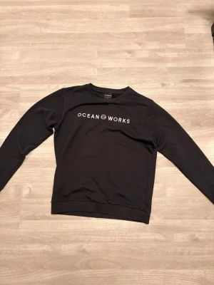 Svart sweatshirt från Ocean Works - Svart sweatshirt från Ocean Works med vit logotyp på bröstet. Tröjan har rund hals, långa ärmar och ribbade muddar vid ärmslut och nederkant. Materialet känns mjukt och bekvämt, perfekt för chill dagar. Enkel och stilren design som funkar till jeans.