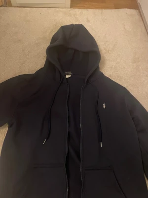 Mörkblå hoodie från Polo Ralph Lauren - Snygg mörkblå hoodie från Polo Ralph Lauren med klassisk logga broderad på bröstet. Tröjan har huva med snörning, dragkedja och två fickor framtill. Perfekt för chill dagar och enkel att matcha med jeans eller joggers. I storlek L fast passar perfekt för dig om du är 175 ungefär