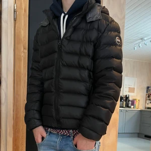 Svart colmar jacka  - Svart pufferjacka från Colmar med huva och dragkedja framtill. Jackan har ett quiltat mönster och logotyp på ärmen. Perfekt för kalla dagar, med en snygg och modern look. Passformen är normal och jackan är tillverkad i syntetmaterial.