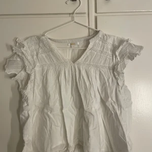 Vit blus med volangärmar - Säljer en söt vit blus från Gina med v-ringning och volangdetaljer på de ärmlösa axlarna. Blusen har fina broderade partier upptill och ett luftigt, lätt tyg som ger en romantisk vibe. Perfekt för dig som gillar boho-stil och ljusa färger.