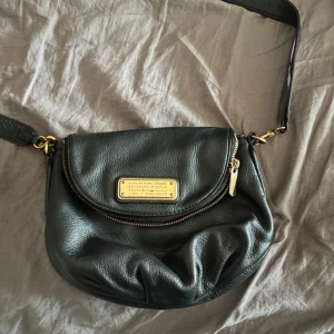 Svart axelväska från Marc Jacobs - Snygg svart axelväska från Marc Jacobs i mjukt skinn med guldfärgade metalldetaljer. Väskan har en rundad form, dragkedja och en metallplatta med logga framtill. Perfekt storlek för det viktigaste och har justerbar axelrem. En skada på ena axelremmens fäste se bild två