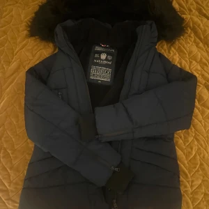 Mörkblå vinterjacka från Navahoo - Snygg mörkblå vinterjacka från Navahoo med fluffig huva och quiltad design. Jackan har dragkedjor framtill, ribbade muddar vid ärmslut och mjukt foder på insidan. Perfekt för kalla dagar och har en modern, figurnära passform. 