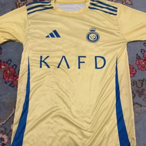 Al Nassr Ronaldo 7 gul fotbollströja - Gul fotbollströja från Al Nassr med blå detaljer och tryck. Adidas-logga på bröstet, klubbmärke och sponsortryck framtill. På ryggen står det Ronaldo och nummer 7 i blått. Tröjan är kortärmad och tillverkad i lätt, ventilerande material.
