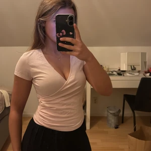 Ljusrosa omlottopp med v-ringning - Säljer en ljusrosa omlottopp med v-ringning och korta ärmar. Toppen har en figurnära passform och är tillverkad i mjuk bomull. Snygg omlottdetalj framtill som ger en feminin look. Perfekt att styla med jeans eller kjol. Helt oanvänd!nypris 259kr