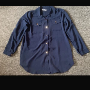 Mörkblå skjorta med fickor - Stilren mörkblå skjorta från Alyra med klassisk krage, två bröstfickor med lock och guldiga knappar framtill. Skjortan har lång ärm och en avslappnad passform, perfekt till jeans eller kjol. Materialet känns mjukt och lätt.