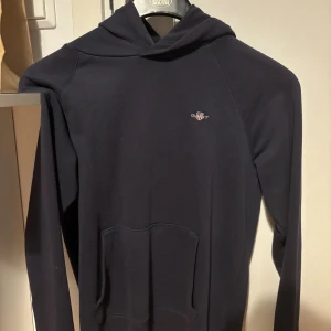 Mörkblå hoodie från Gant - Snygg mörkblå hoodie från Gant med liten broderad logga på bröstet. Klassisk känguruficka framtill och huva. Perfekt för dig som gillar stilrena och bekväma plagg. Passar dig som vill ha en enkel men ändå exklusiv look.