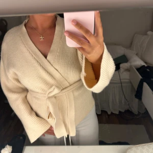 Beige omlott kofta med bälte - Säljer en beige ribbstickad omlottkofta med bälte i midjan. Koftan har långärmade, lite vidare ärmar och är gjord i ett mjukt stickat material som känns riktigt skönt mot huden. Perfekt för dig som gillar en chill och stilren look.