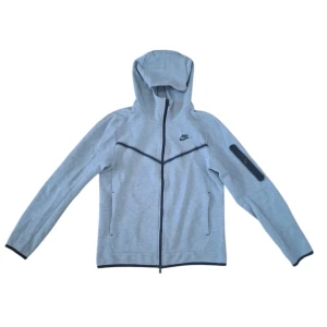 Grå vindjacka med huva från Nike - Nike vindjacka i ljusgrått med svart logga på bröstet och svarta detaljer. Jackan har hel dragkedja, huva och lång ärm samt en praktisk ficka med dragkedja på ena ärmen. Perfekt för dig som gillar sportig och clean stil.