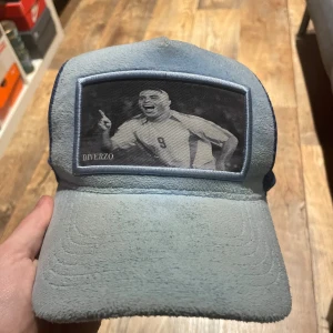 Blå keps från Diverzo Limited Edition - Snygg blå keps från Diverzo Limited Edition med mesh på sidorna och baksidan. Framsidan har en patch med en svartvitt bild på Ronaldo Nazario. Kepsen har böjd skärm och justerbar snapback. Perfekt för dig som gillar sportig stil.