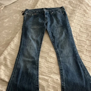 True Religion bootcut jeans blå - Snygga blå jeans från True Religion med klassisk bootcut-modell och ikonisk broderad bakficka. Jeansen har låg midja, silverfärgade knappar och detaljerade sömmar. 
