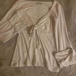 Vit omlottopp med knyt från Design by Si - Supersöt vit omlottopp från Design by Si i mjuk bomull. Toppen har långärmade ärmar med lätt volym och snygg v-ringning framtill. 