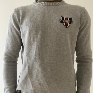 Grå stickad tröja från Morris - Snygg grå stickad tröja från Morris med broderad logga på bröstet. Tröjan har rund hals, ribbade muddar vid ärmslut och nederkant samt långärmad passform. Perfekt för dig som gillar stilrena och klassiska plagg med en sportig touch.