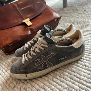 Blå sneakers från Premiata - Snygga blå sneakers från Premiata med beige detaljer och vit häl. Skorna har en rund tå, snörning och är tillverkade i mocka med gummisula. Premiata-logga på sidan och bak på hälen. Perfekt för dig som gillar stilrena och klassiska sneakers. Kom med bud!