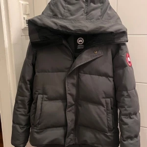 Grå pufferjacka från Canada Goose - Säljer en grå pufferjacka från Canada Goose med stor huva och klassisk logga på ärmen. Jackan har dragkedja och tryckknappar framtill, samt två stora fickor. Perfekt för kalla dagar och har en clean, modern look.
