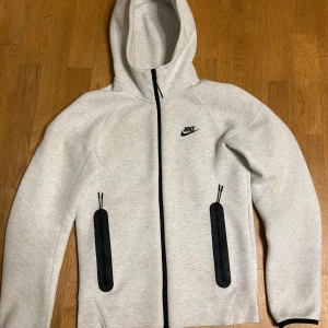 Nike tech - Nike hoodie i ljusgrått med svarta dragkedjor på fickorna och svart Nike-logga på bröstet. Tröjan har hel dragkedja framtill, huva och långa ärmar. Perfekt för chill eller träning, riktigt clean och sportig vibe.