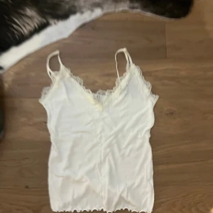 Vit linnetopp med spetsdetaljer - Säljer en söt vit linnetopp med smala axelband och v-ringad hals. Toppen har fina spetsdetaljer längs halsringningen och nederkanten, vilket ger en romantisk vibe. Perfekt för att styla med jeans eller kjol för en chill look.