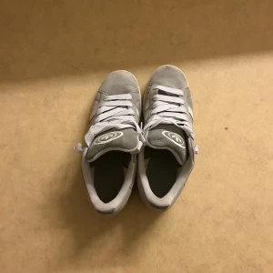 Grå Adidas Campus sneakers mocka - Säljer ett par grå Adidas Campus sneakers i mocka med vita detaljer och klassiska tre ränder på sidan. Skorna har rund tå, platt sula och vita snören. Adidas-logga på plösen och stilren design som passar till jeans eller shorts. Har ej använt de mycket och priset går att diskuteras