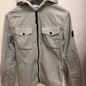 Beige vindjacka från Stone Island - Snygg beige vindjacka från Stone Island med huva och två stora bröstfickor med knappar. Jackan har svart dragkedja framtill och klassisk Stone Island patch på vänster ärm. Perfekt för dig som gillar streetwear och vill ha en clean look.