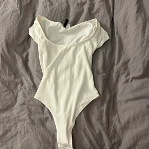 Vit offshoulder bodysuit från H&M - Säljer en vit bodysuit från H&M Divided i storlek XS. Modellen har offshoulder och korta ärmar, med en snygg och enkel design som sitter tight mot kroppen. Materialet är mjuk bomull som känns skönt mot huden. Perfekt för att skapa en clean och fräsch look.