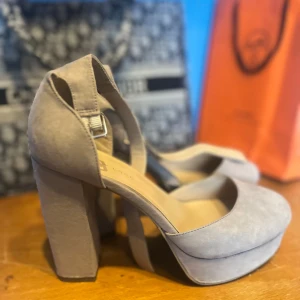 Beige mocka pumps från New Look - Säljer ett par snygga pumps från New Look i beige mocka med rund tå och hög klack. Skorna har en bred plattform och ett justerbart spänne vid ankeln. Perfekta för dig som gillar chunky heels och vill sticka ut lite extra.