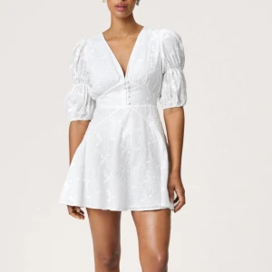 Vit kort klänning med puffärm - Supersöt vit kort klänning med puffiga kortärmar och v-ringad hals. Klänningen har broderade blommor, knappar framtill och markerad midja som ger en snygg siluett. Perfekt till student eller sommaren