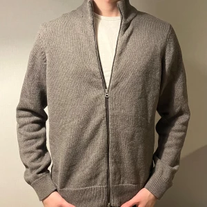 Grå stickad kofta från Dobber - Snygg grå stickad kofta från Dobber i storlek medium. Koftan har hel dragkedja framtill, ribbade muddar och en hög krage. Perfekt för lager-på-lager och chill stil. Enkel design utan mönster, passar dig som gillar cleana plagg.