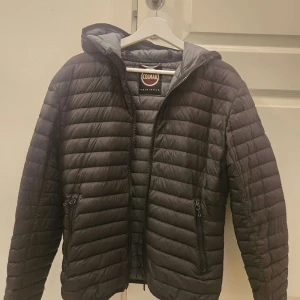 Svart dunjacka från Colmar - Snygg svart dunjacka från Colmar med huva och horisontella quiltade sömmar. Jackan har dragkedja framtill, två fickor med dragkedja och Colmar-logga på bröstet. Har ett litet hål på armen (se sista bilden) men hålet är lagat.