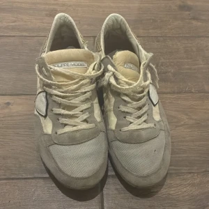 Beige sneakers från Philippe Model - Snygga sneakers från Philippe Model i beige och grå toner. Skorna har ovandel i mocka och textil, med klassisk snörning och gummisula. Ikonisk sköld-detalj på sidan och diskret logga på plösen. Perfekta för en avslappnad och trendig look.