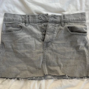 Grå jeanskjol från 157, storlek M - Grå jeanskjol från 157 i klassisk femficksmodell med rå, fransig kant nertill. Kjolen har knappar framtill, bälteshällor och bakfickor. Perfekt för dig som gillar en avslappnad och trendig denimlook.