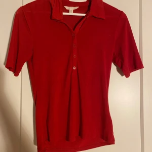 Röd kortärmad t shirt med krage - Säljer en röd t-shirt från H&M i storlek S. Toppen har klassisk krage, knappar framtill och korta ärmar. Materialet är mjukt och lätt, perfekt för dig som gillar enkel och stilren design. Passformen är normal och färgen är riktigt snyggt röd.