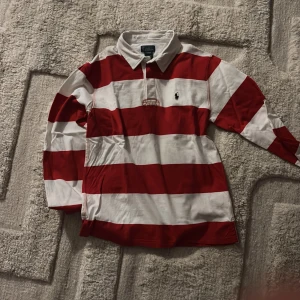 Randig röd/vit pikétröja Polo Ralph Lauren - Säljer en klassisk pikétröja från Polo Ralph Lauren i storlek L. Tröjan har breda röda och vita ränder, lång ärm och krage. Ikoniska Polo-loggan broderad på bröstet. Perfekt för dig som gillar sportig och preppy stil.