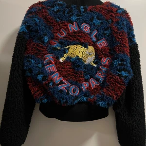 Kenzo x H&M teddyjacka med tiger - Unik teddyjacka från Kenzo x H&M i svart, blått och rött med broderad tiger och texten 'Jungle Kenzo Paris' på ryggen. Jackan har långärmade, croppad passform och knappar framtill. Materialet är en mix av akryl, modakryl och polyester.
