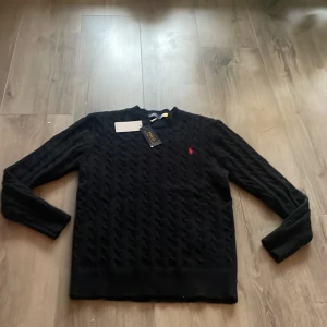 Svart kabelstickad tröja Polo Ralph Lauren - Svart kabelstickad tröja från Polo Ralph Lauren med klassisk röd broderad logga på bröstet. Tröjan har rund halsringning och långa ärmar, perfekt för lager-på-lager. Snygg och tidlös design som funkar till allt.