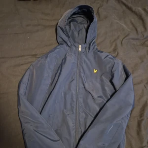 Marinblå windbreaker Lyle & Scott Junior - Snygg marinblå windbreaker från Lyle & Scott Junior med luva och gul logga på bröstet. Det gröna syns inte när man har på sig den. Passar dig som är 155-175cm lång. Perfekt för dig som vill ha en stilren jacka.