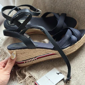 REA Svarta sandaler med kilklack Tommy Hilfiger - Snygga svarta sandaler från Tommy Hilfiger med flätad kilklack i naturfärgat material och dekorativt band med logga på sidan. Remmarna är i skinn och textil, och sandalerna har spänne runt ankeln för extra support. Perfekta för sommarens alla tillfällen.