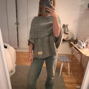 Grå stickad ponchotröja cape - Säljer en grå stickad ponchotröja  med bred ribbad krage och kabelstickat mönster. Tröjan har en lös passform och korta ärmar, perfekt för lager på lager under höst och vinter. 